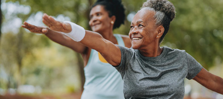 Deux Afro-Américaines septuagénaires sourient en faisant du yoga dans un parc entouré d'arbres.